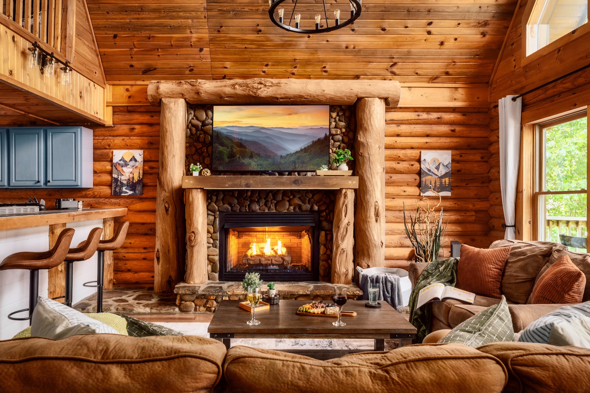 Cabin living room with a stone fireplace, log walls, and a television above the mantel.