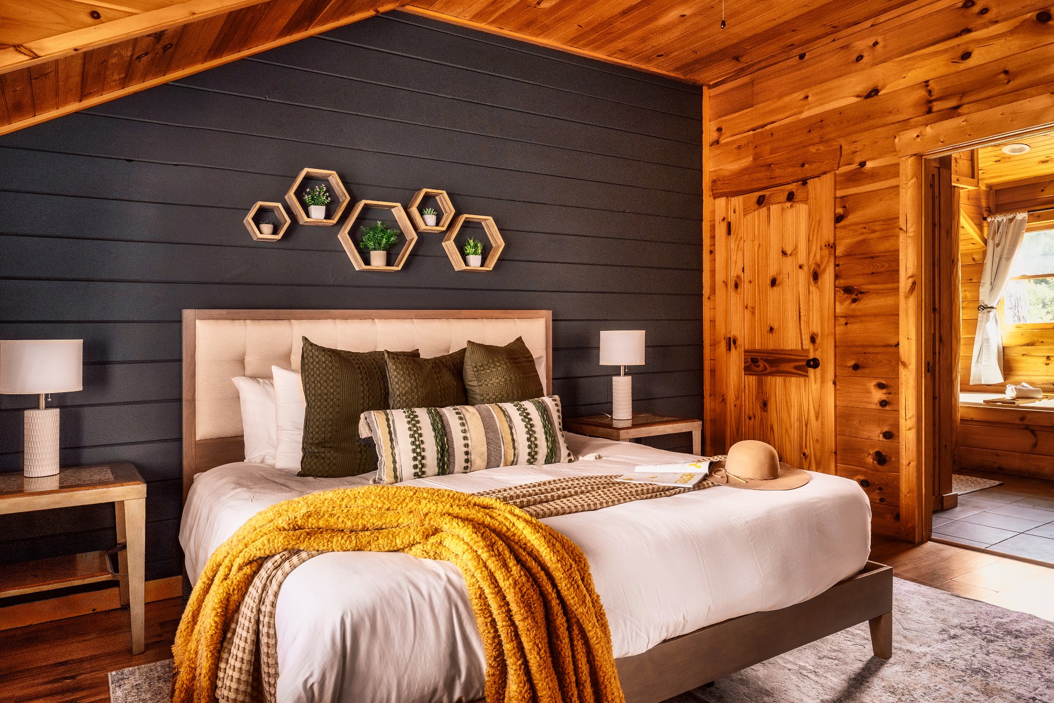 Rustic cabin bedroom with a white bed, mustard throw, charcoal accent wall, and timber ceiling.