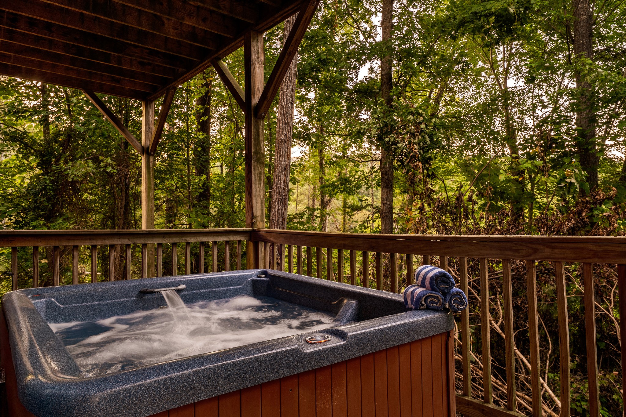 Hot tub on a covered wooden deck overlooking dense trees, with rolled towels beside it.