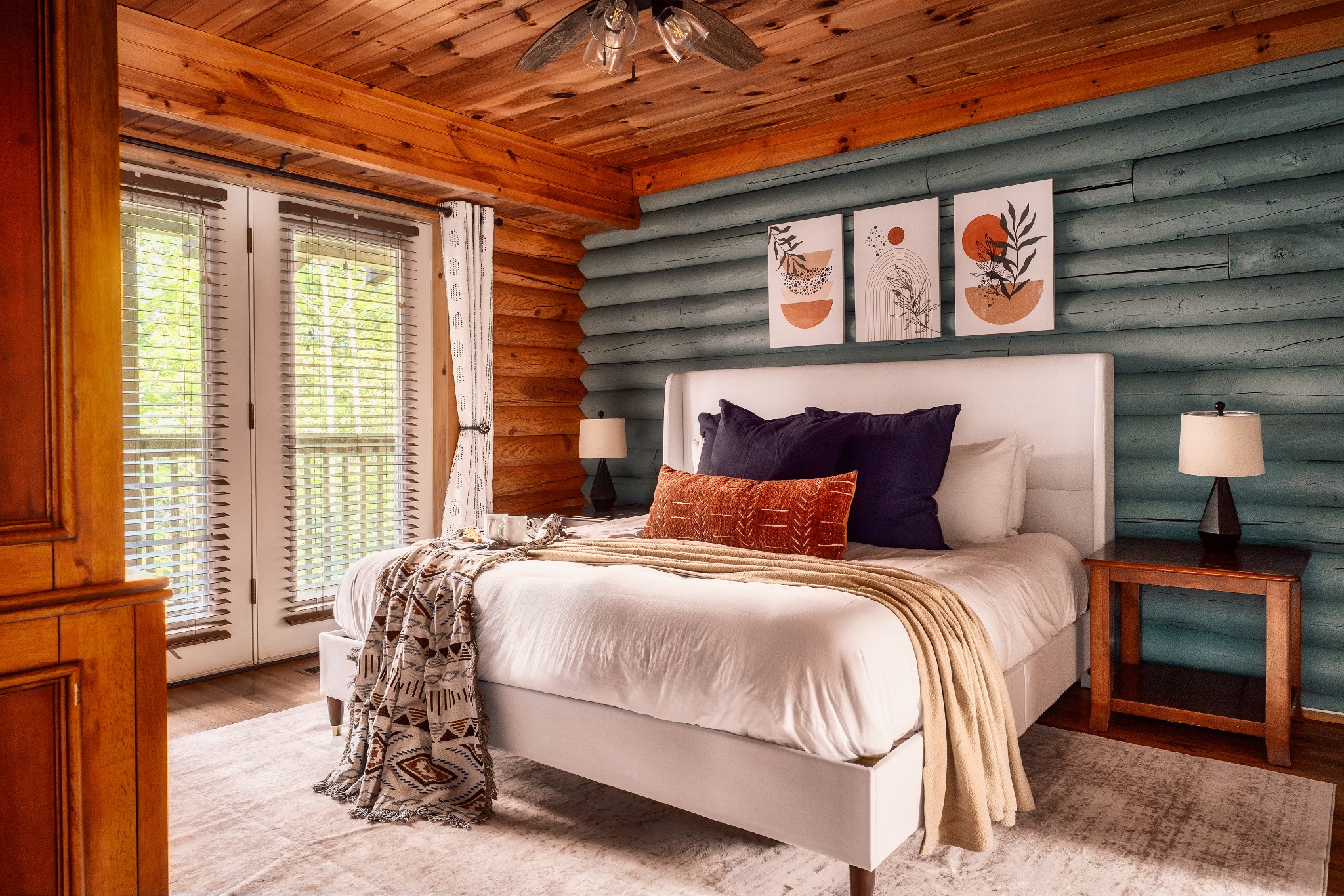 Cabin bedroom with a white bed, teal log walls, warm wood ceiling, and glass doors with blinds.