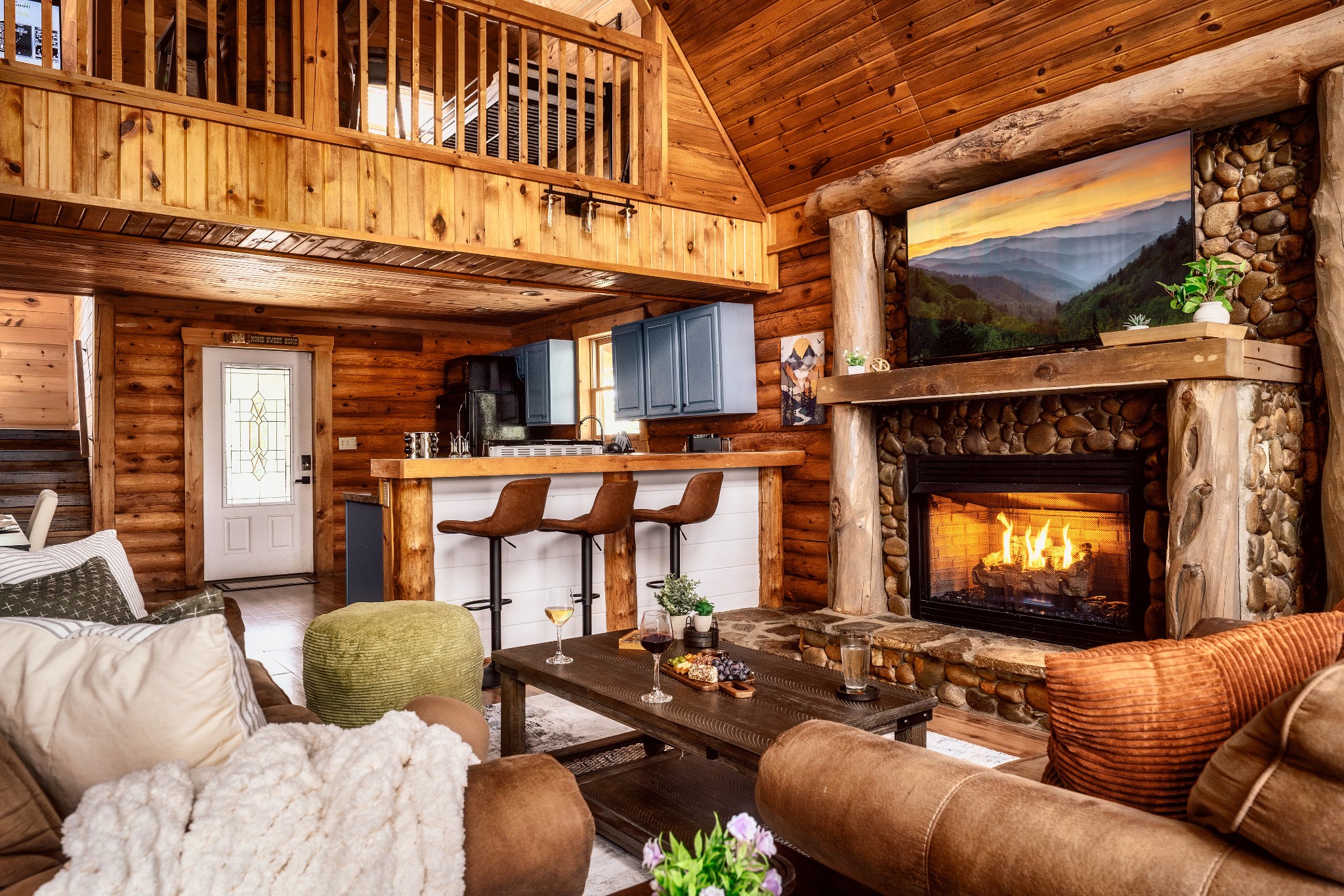 Rustic cabin living room with a stone fireplace, open kitchen, and loft railing.