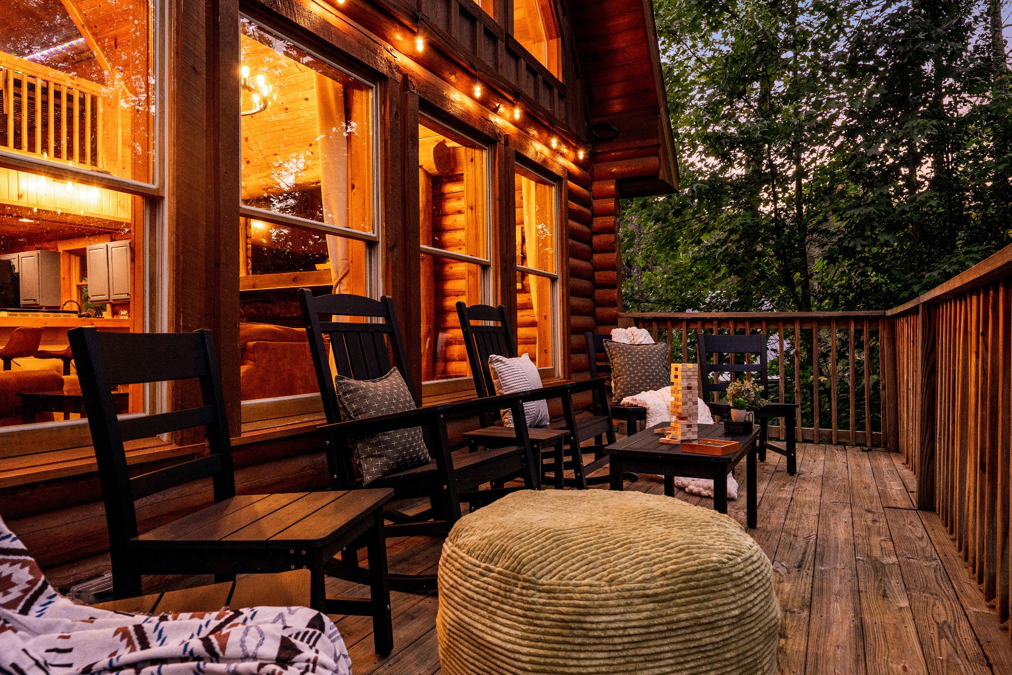 Log cabin deck with Adirondack chairs, a woven ottoman, and warm string lights among trees.