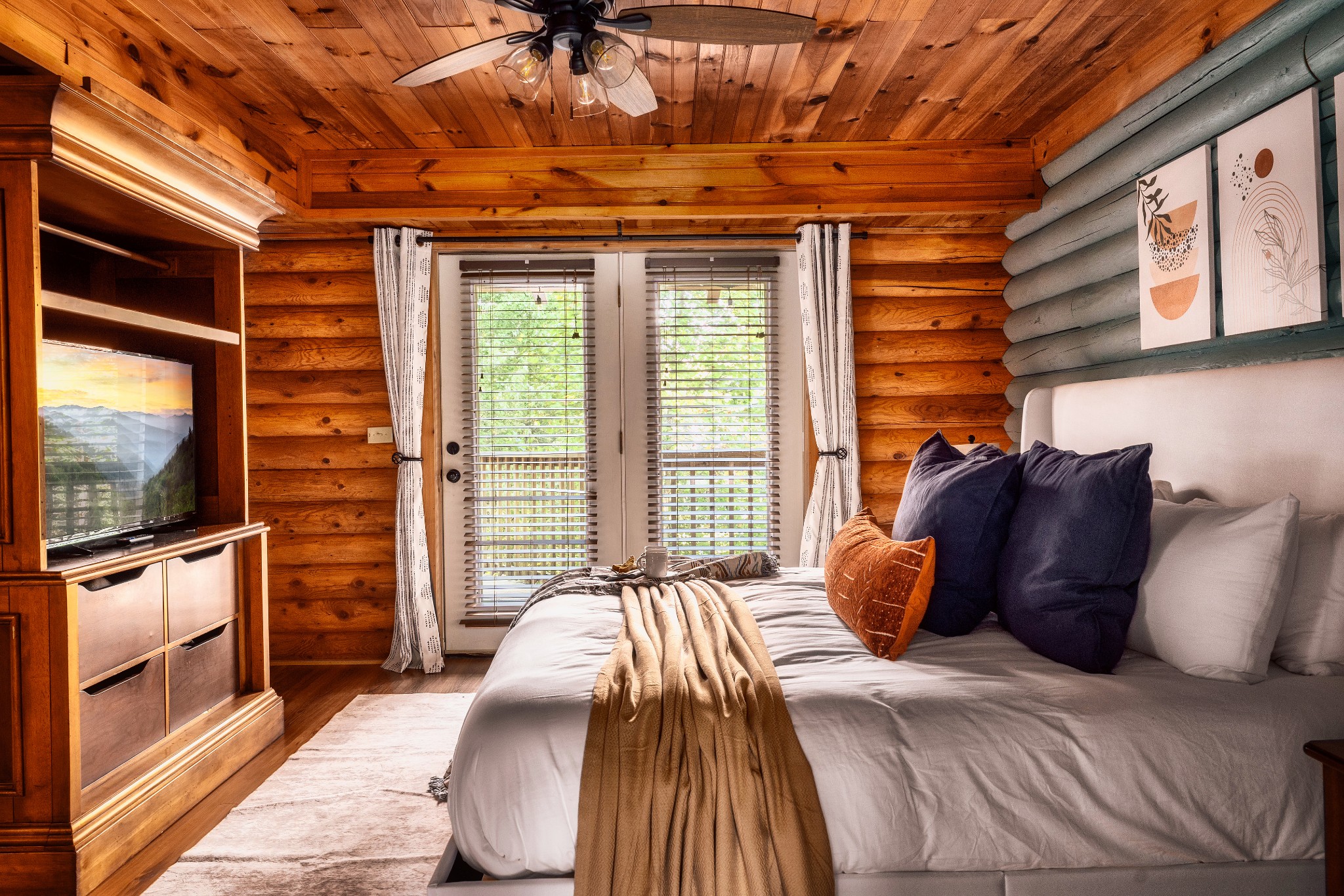 Log cabin bedroom with a white bed, colorful pillows, wood-paneled walls, and double glass doors.