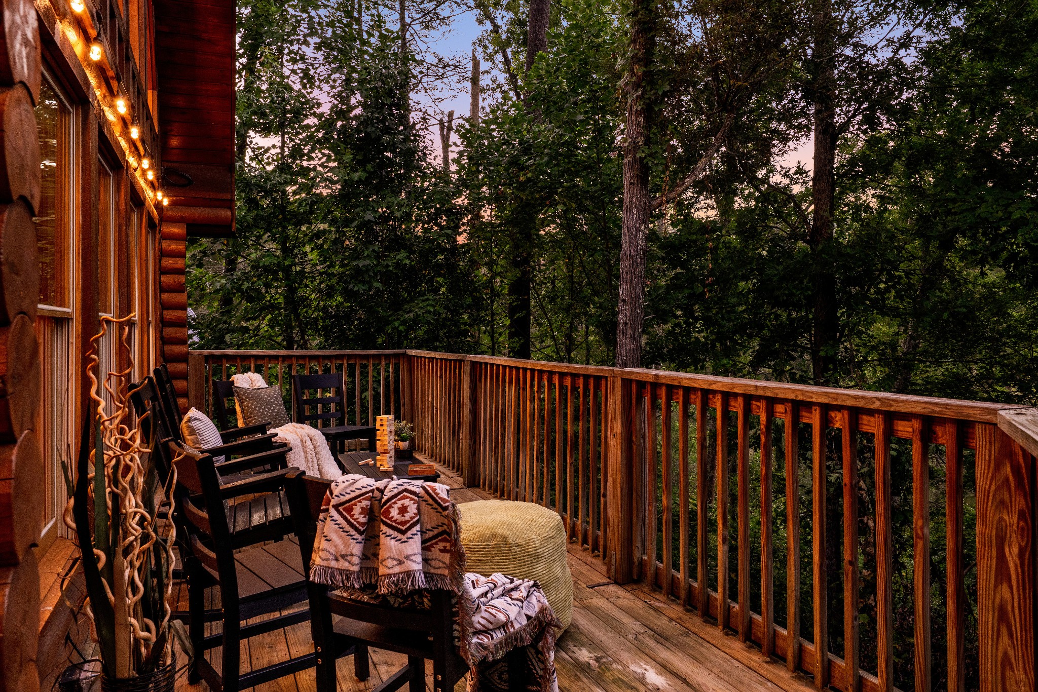 Furnished cabin deck with Adirondack chairs and warm lights overlooking dense woods at dusk.