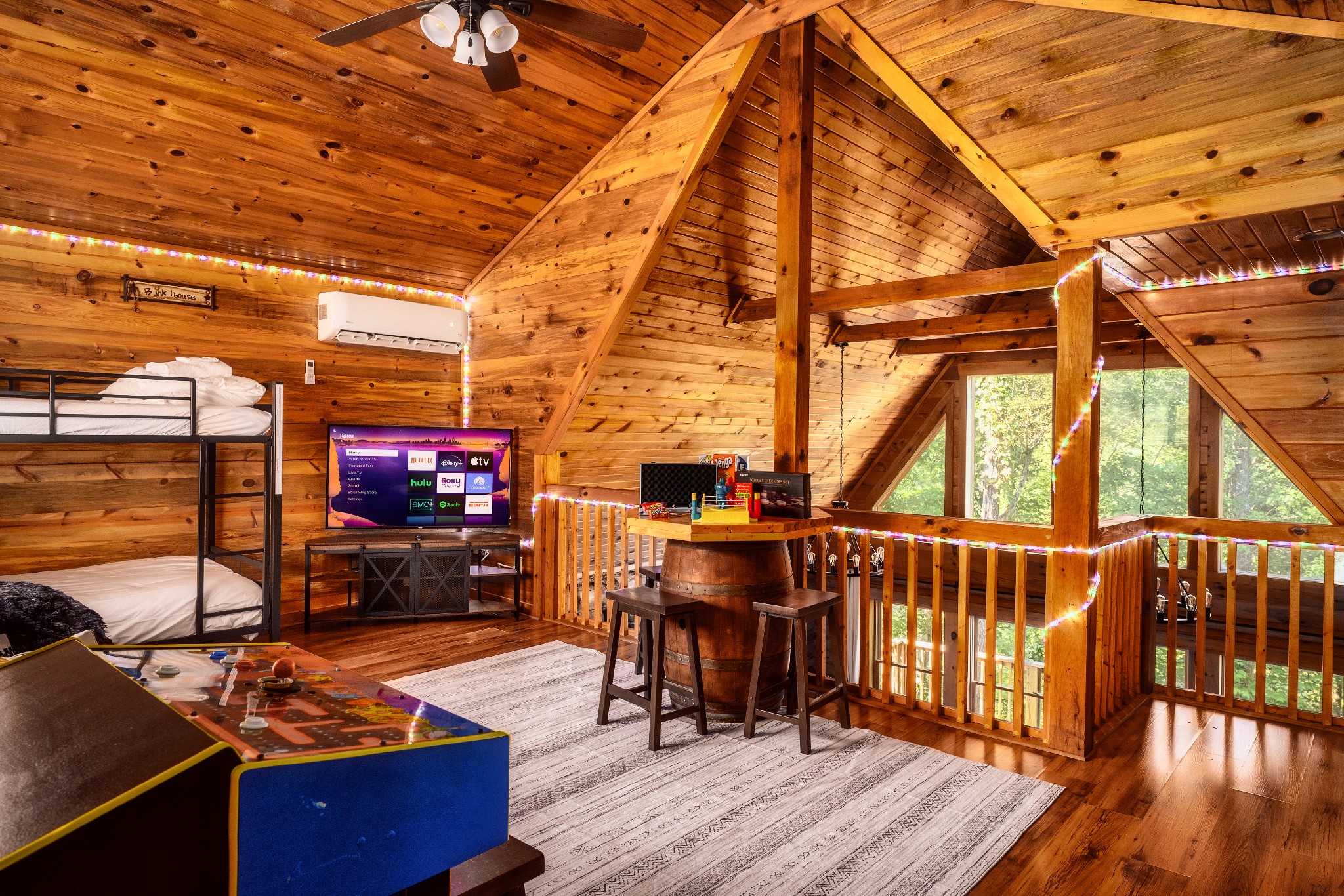 Cabin loft with bunk beds, foosball table, barrel table, and wooded views through large windows.