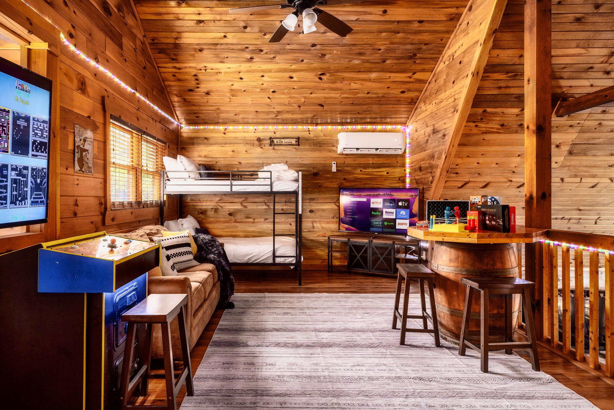Wood-paneled cabin game room with bunk beds, arcade game, television, and barrel bar seating.