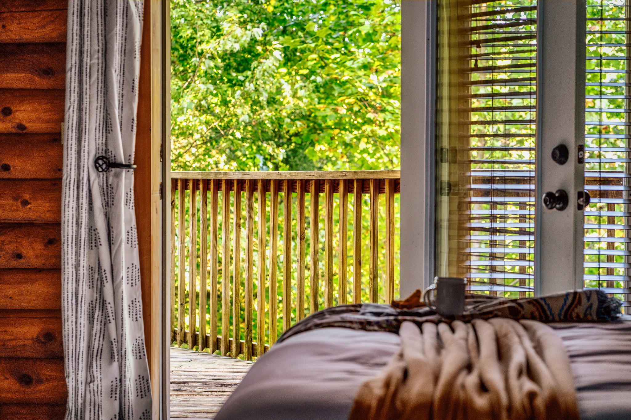 Cabin bedroom with a made bed beside an open door to a leafy wooden balcony.