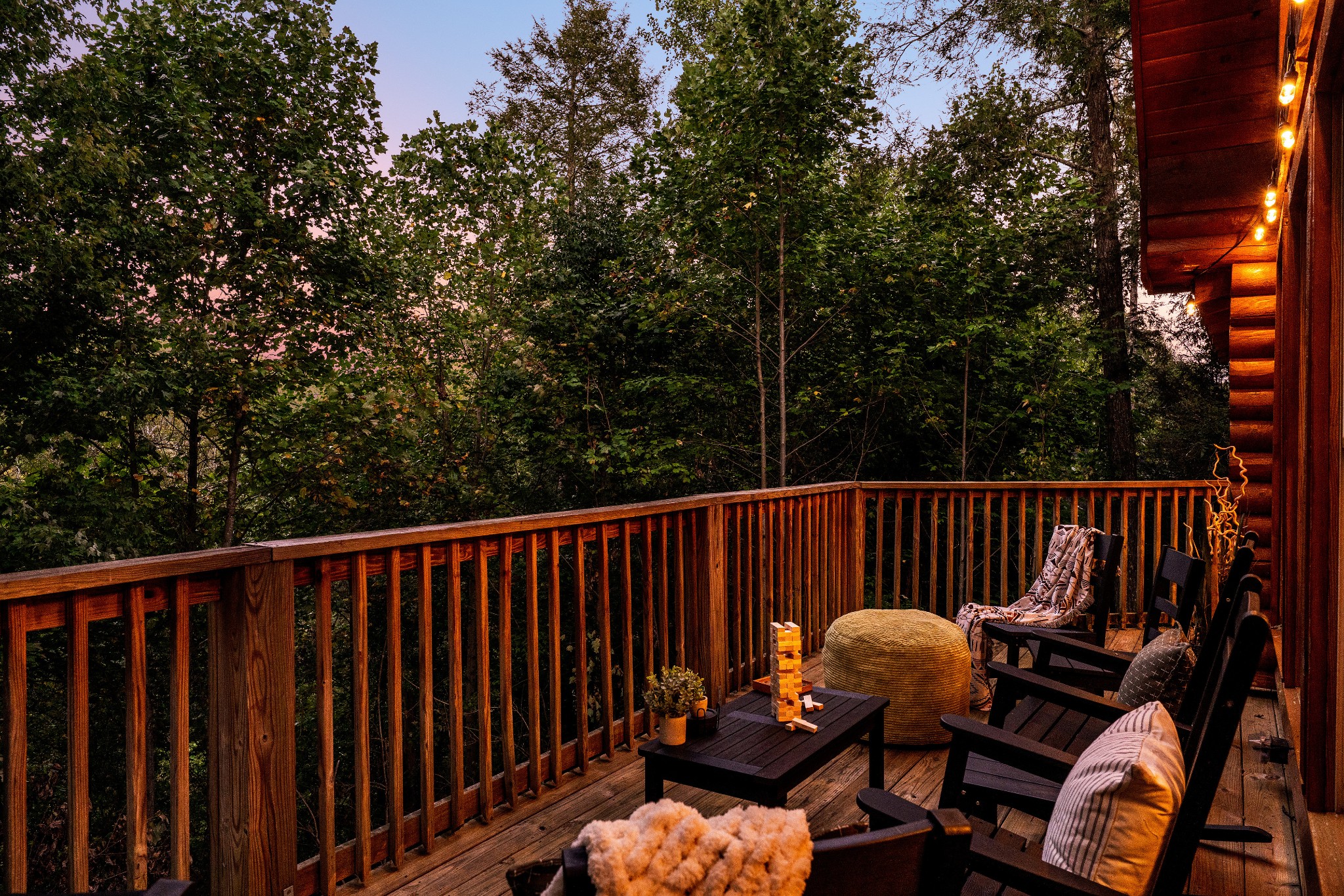 Furnished cabin deck with Adirondack chairs and warm lights, overlooking dense trees at dusk.