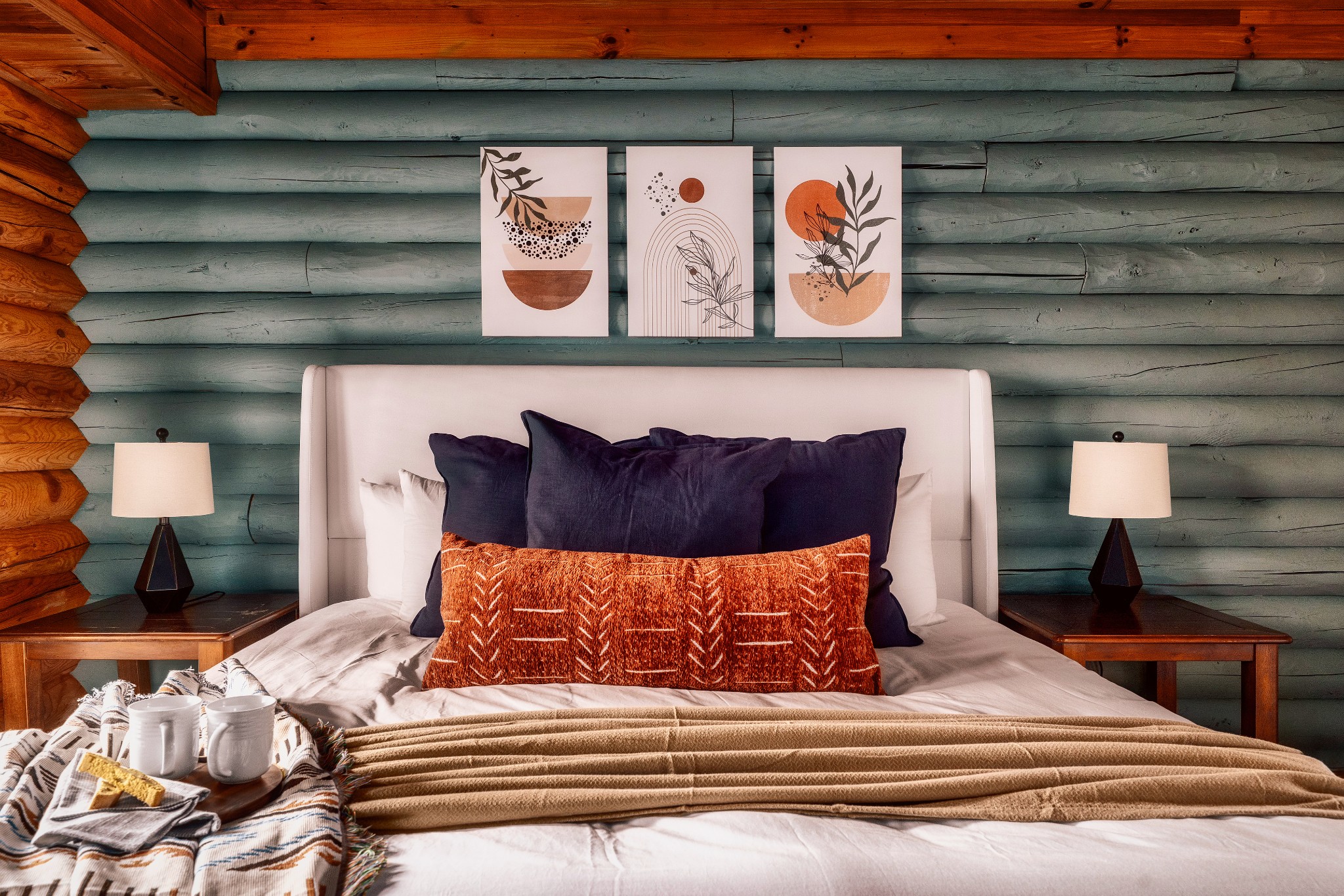Rustic cabin bedroom with teal log walls, a white headboard, navy pillows, and a rust-colored accent pillow.