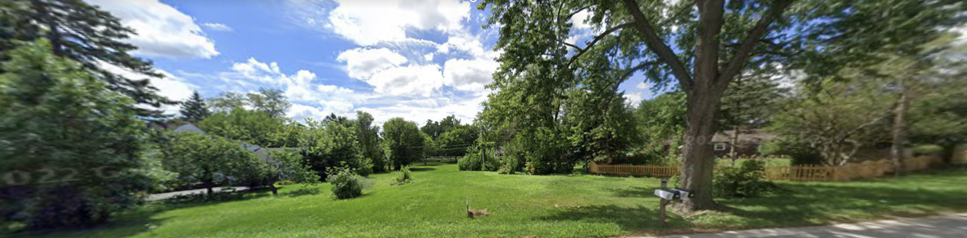 Grassy residential lot with mature trees, neighboring homes, and a roadside mailbox.