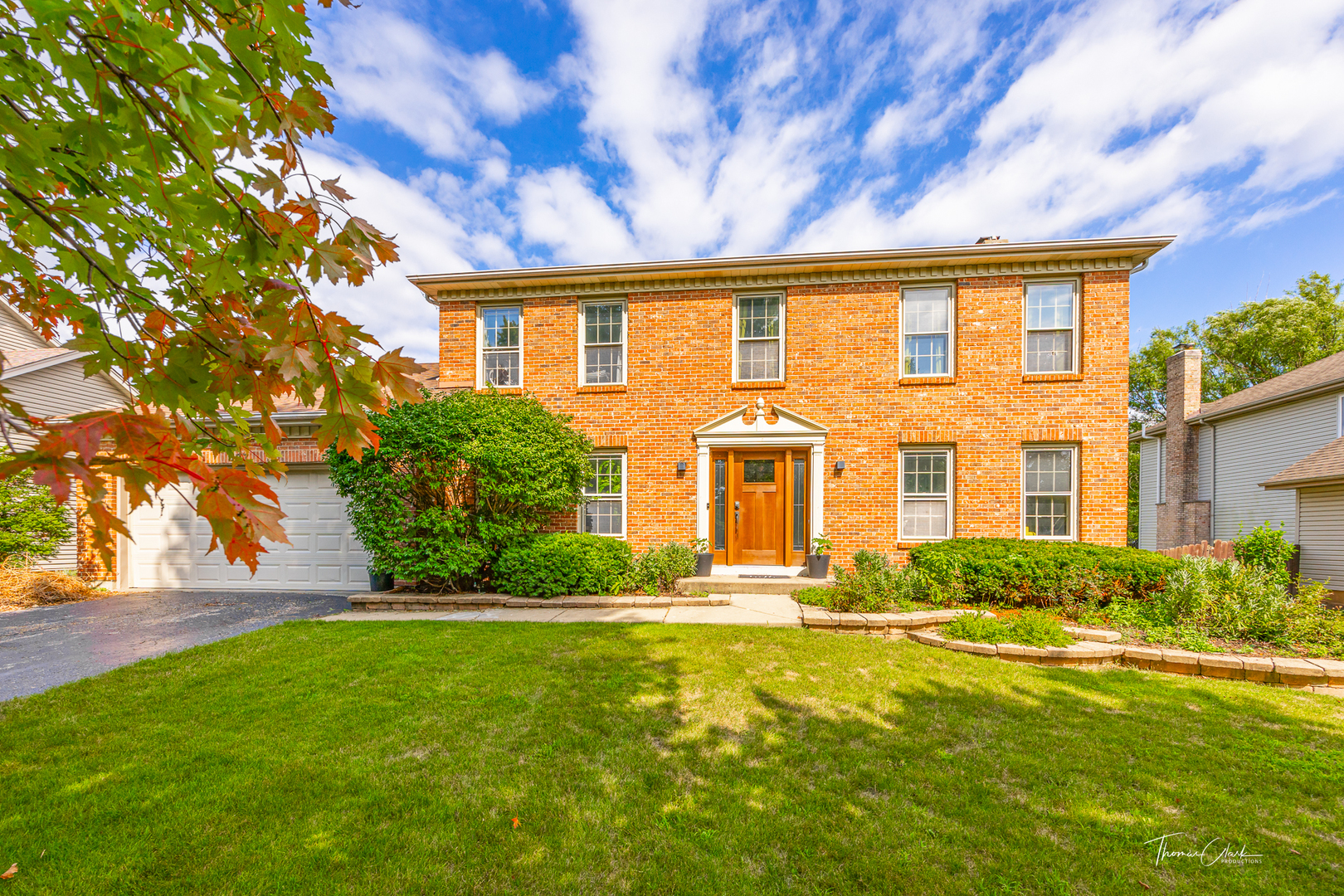 Two-story brick house with an attached garage, wood front door, landscaped shrubs, and a green lawn.