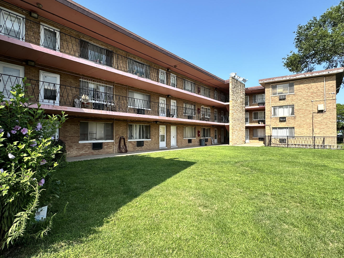 Brick apartment buildings with long balconies surround a sunlit grassy courtyard.