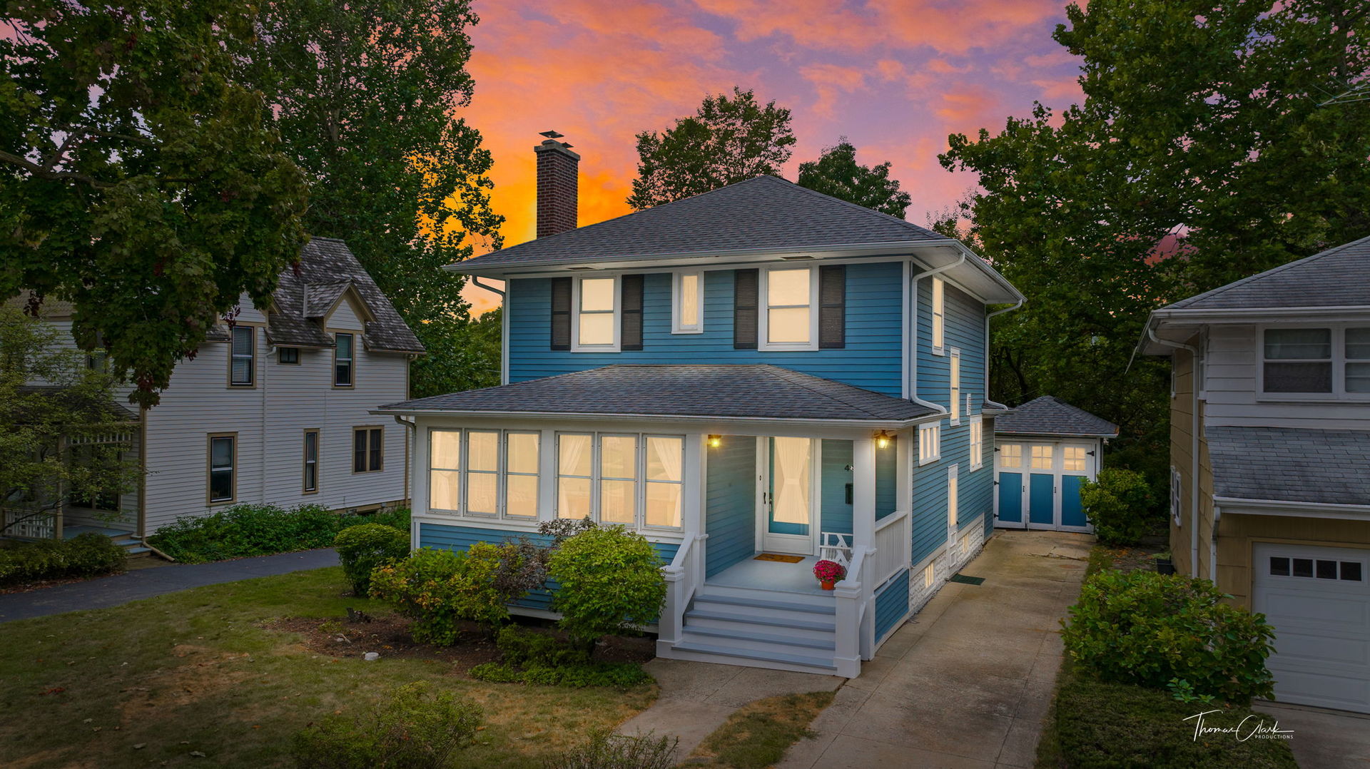 Blue two-story house with a covered porch and detached garage beneath a colorful dusk sky.