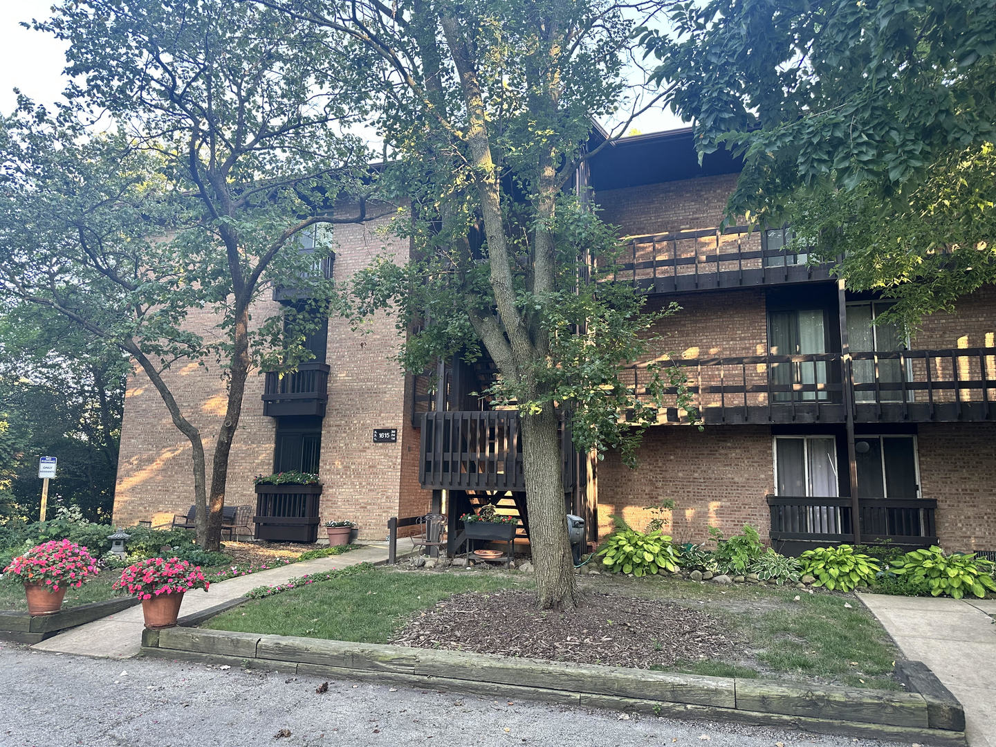 Brick condominium building with dark-railed balconies, mature trees, and landscaped flower beds.