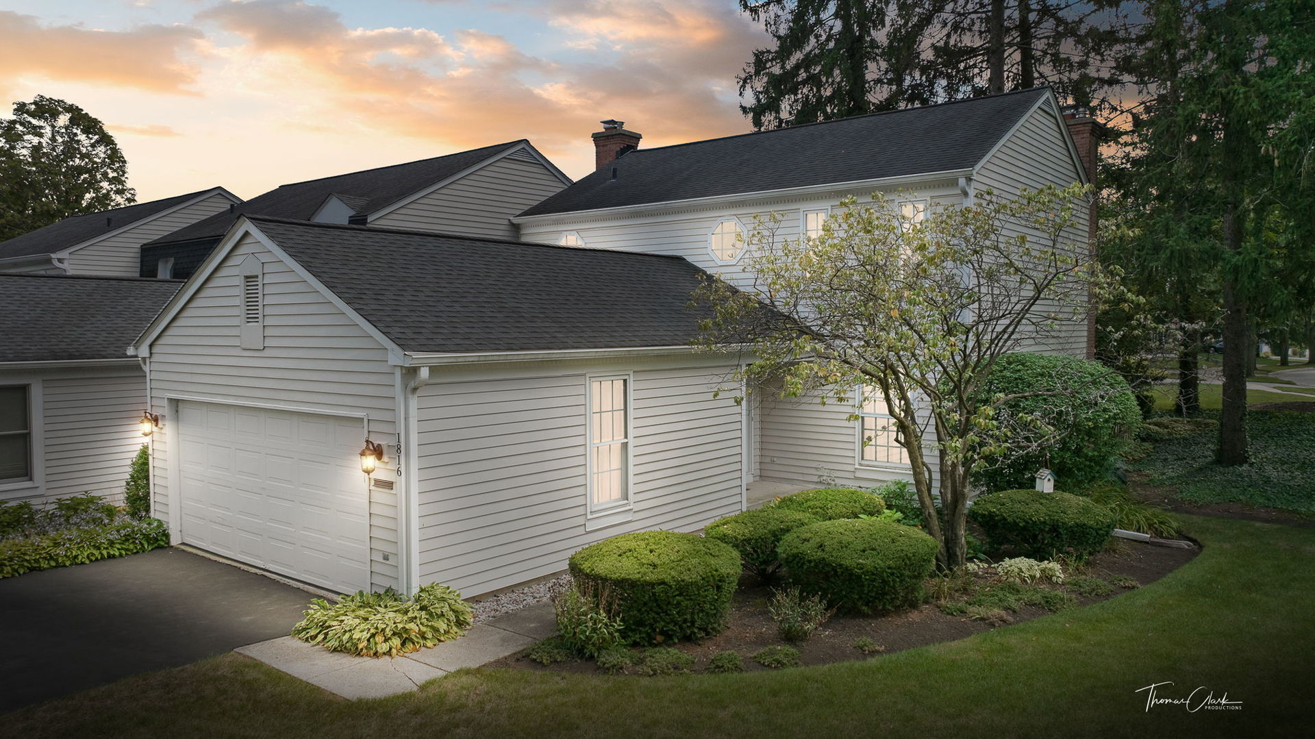 Two-story light-colored house with an attached garage, landscaped shrubs, and mature trees at sunset.