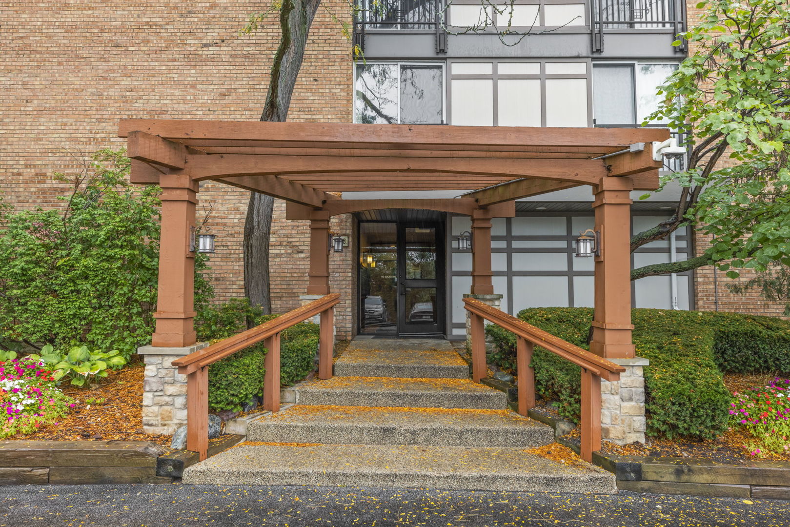Brick condominium entrance beneath a wooden pergola, with steps, ramp, and autumn leaves.