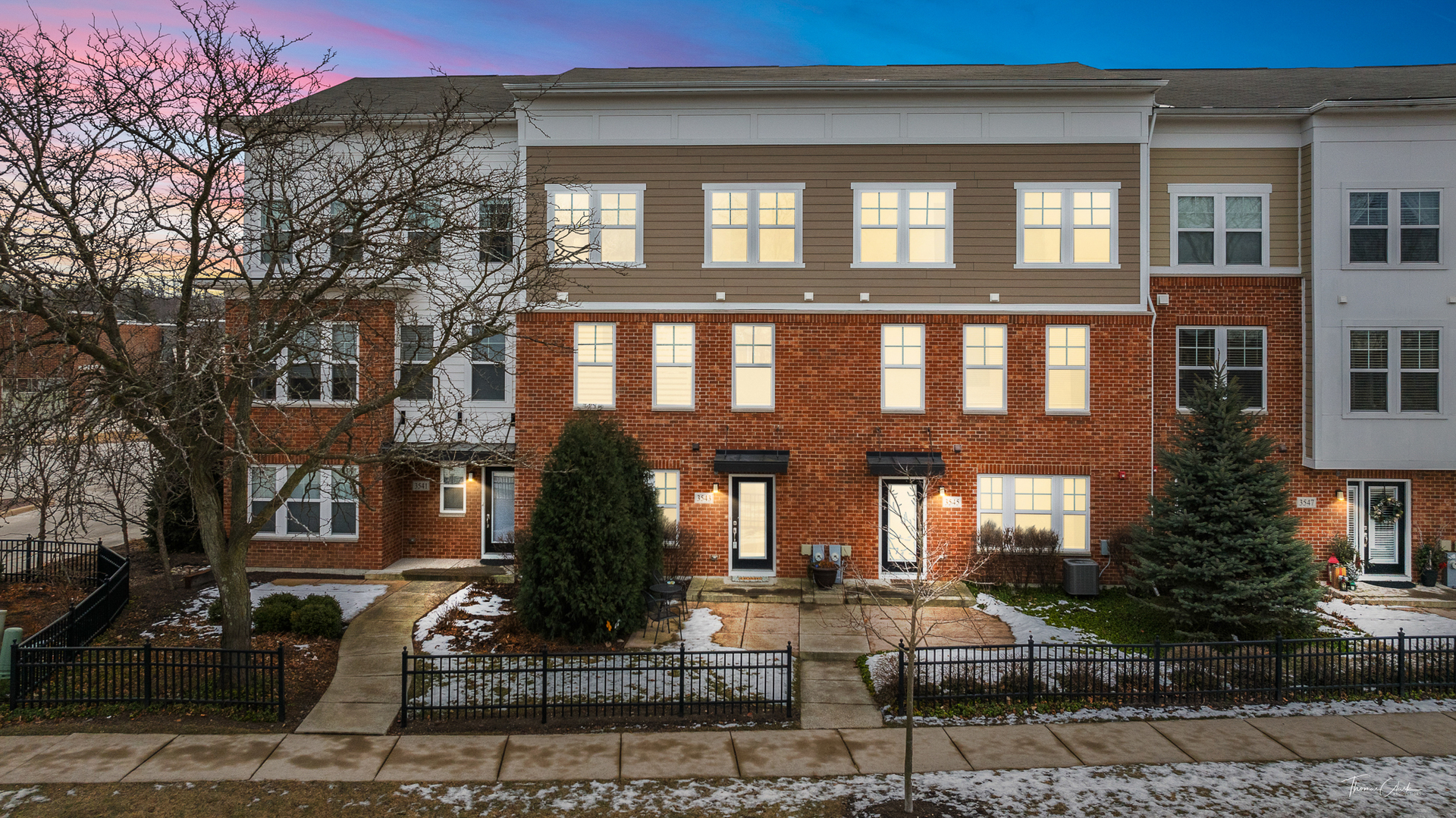 Townhome at 3543 with red brick, brown siding, glowing windows, and snow along the fenced walkway.
