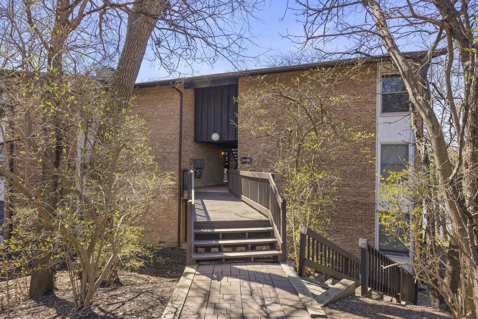 Brick residential building entrance with a raised wooden walkway and steps, framed by budding trees.