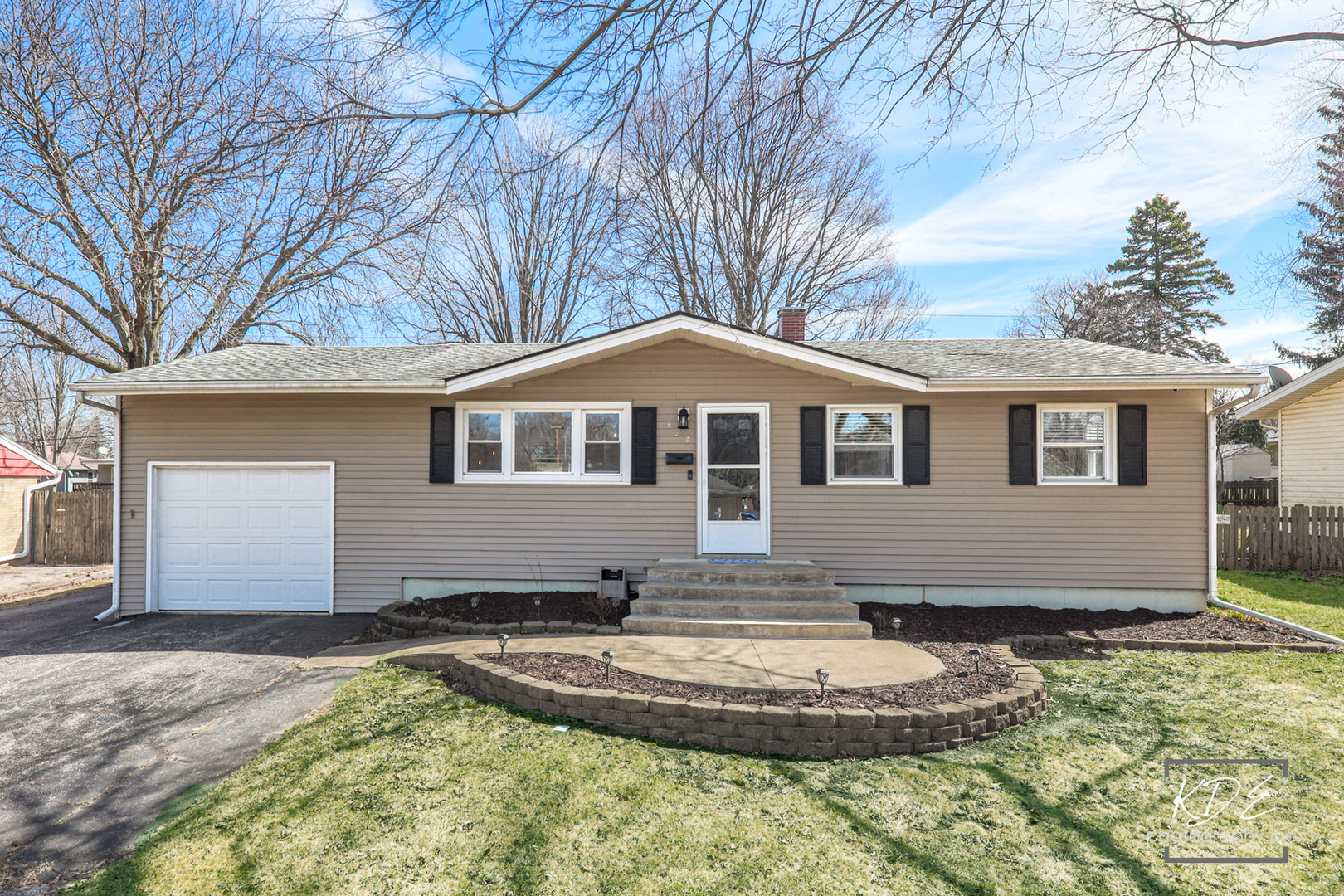Single-story beige ranch house with an attached garage, black shutters, and a curved front walkway.