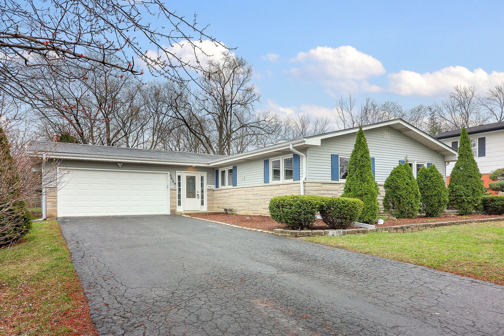 Single-story ranch home with an attached garage, stone facade, blue shutters, and evergreen landscaping.