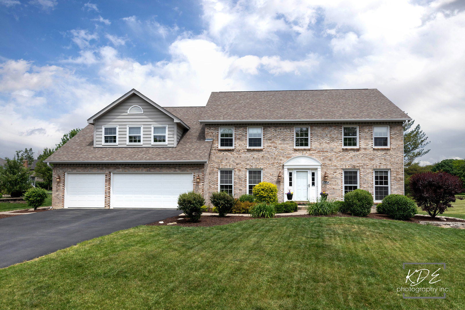 Two-story brick home with an attached two-car garage, landscaped lawn, and wide driveway.