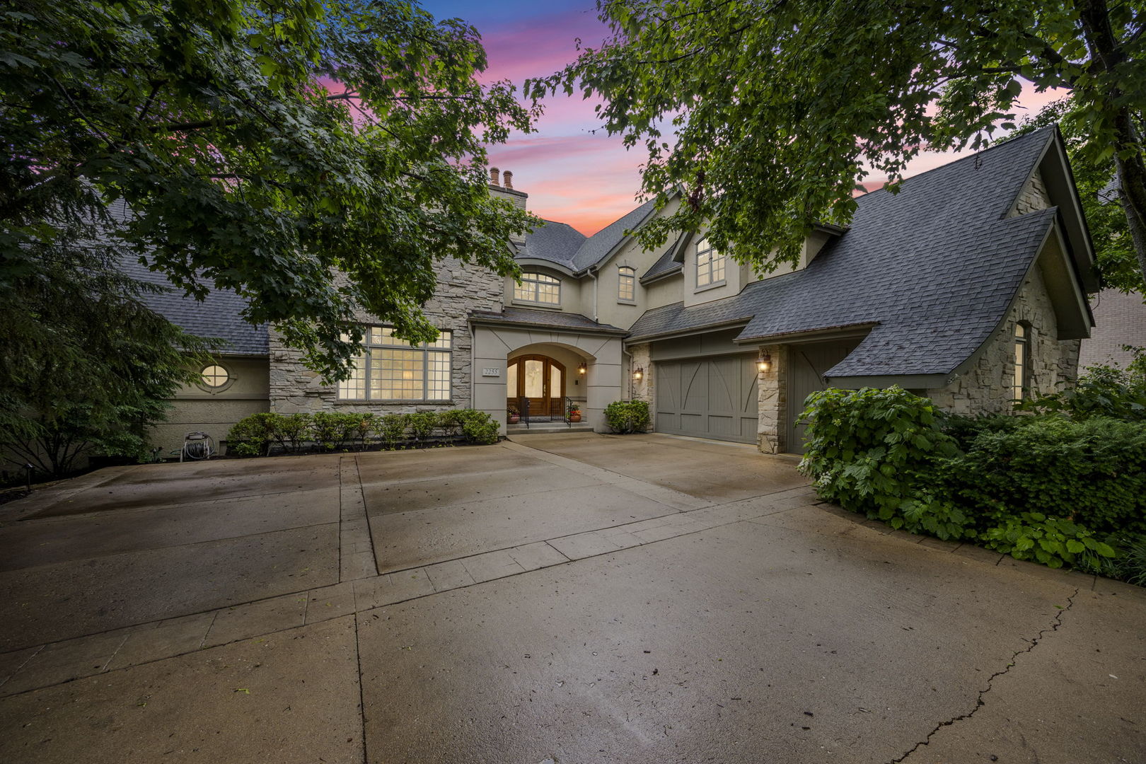 Large stone-and-stucco home with an attached garage and wide driveway beneath a pink dusk sky.