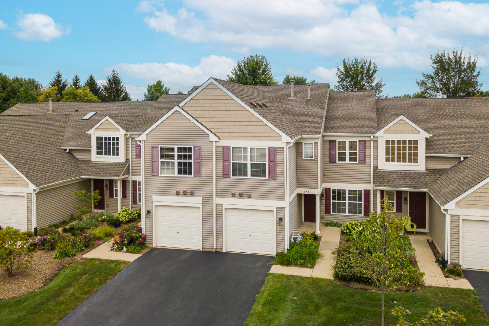 Beige townhome with burgundy shutters, two garage doors, and landscaped flower beds.