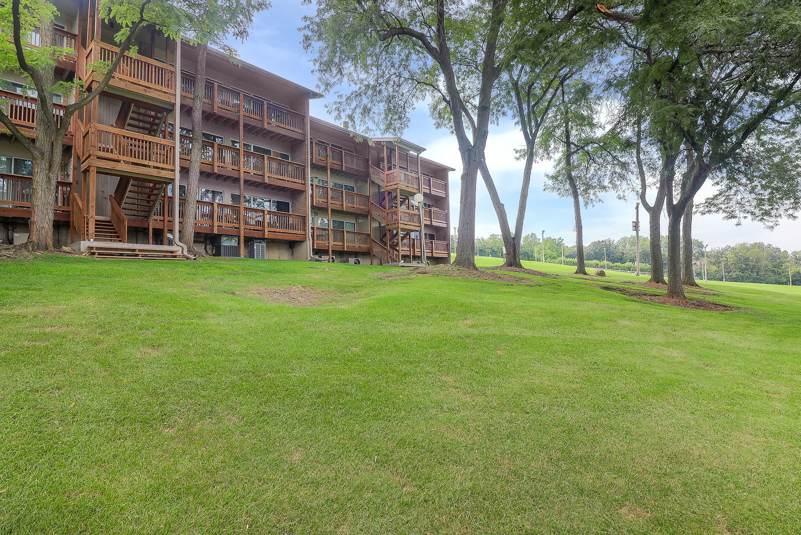 Multi-story condominium with wood balconies and stairways beside a tree-lined lawn.