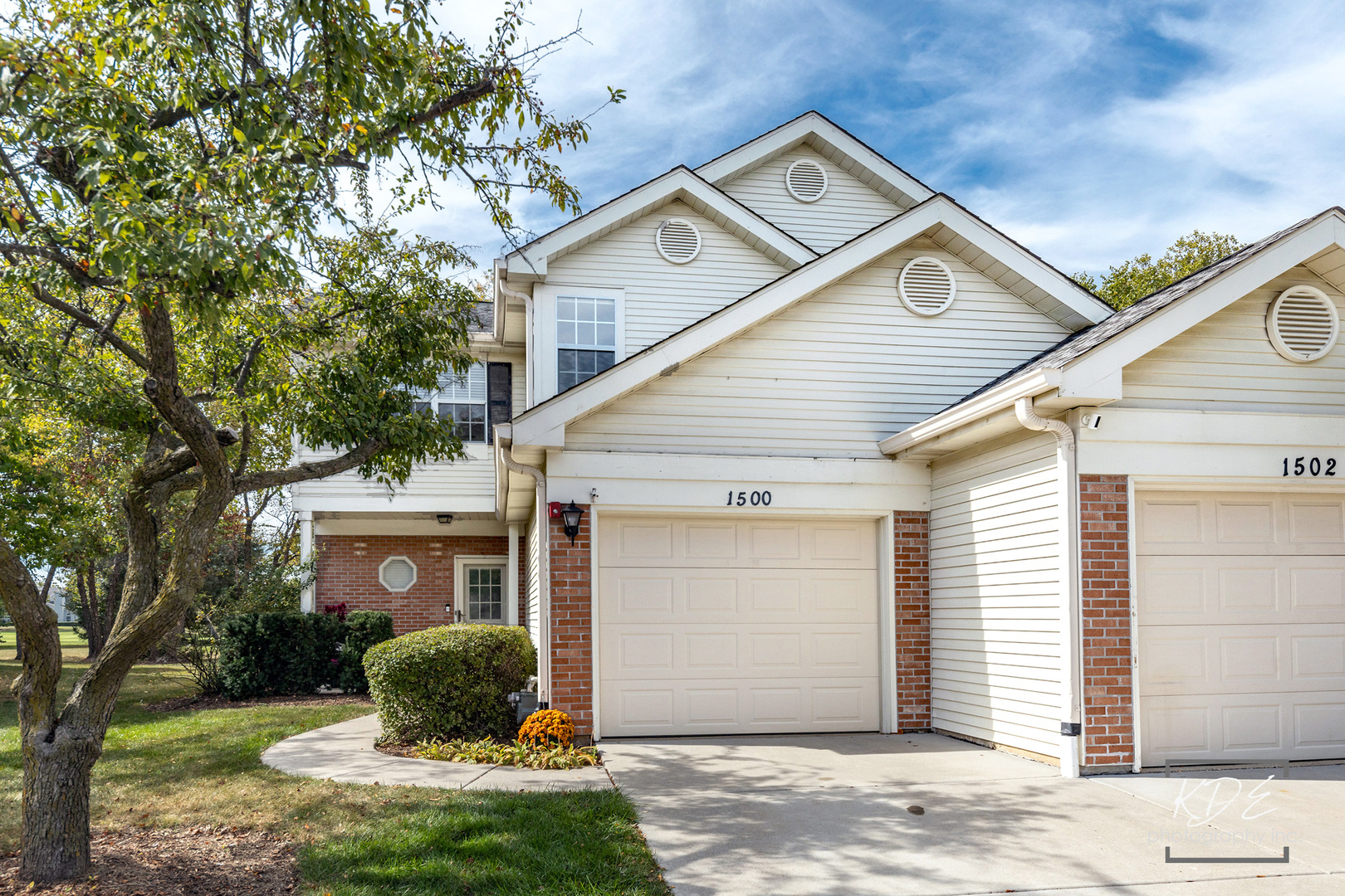 White-sided home with brick accents, two garage doors, and a large tree beside the driveway.