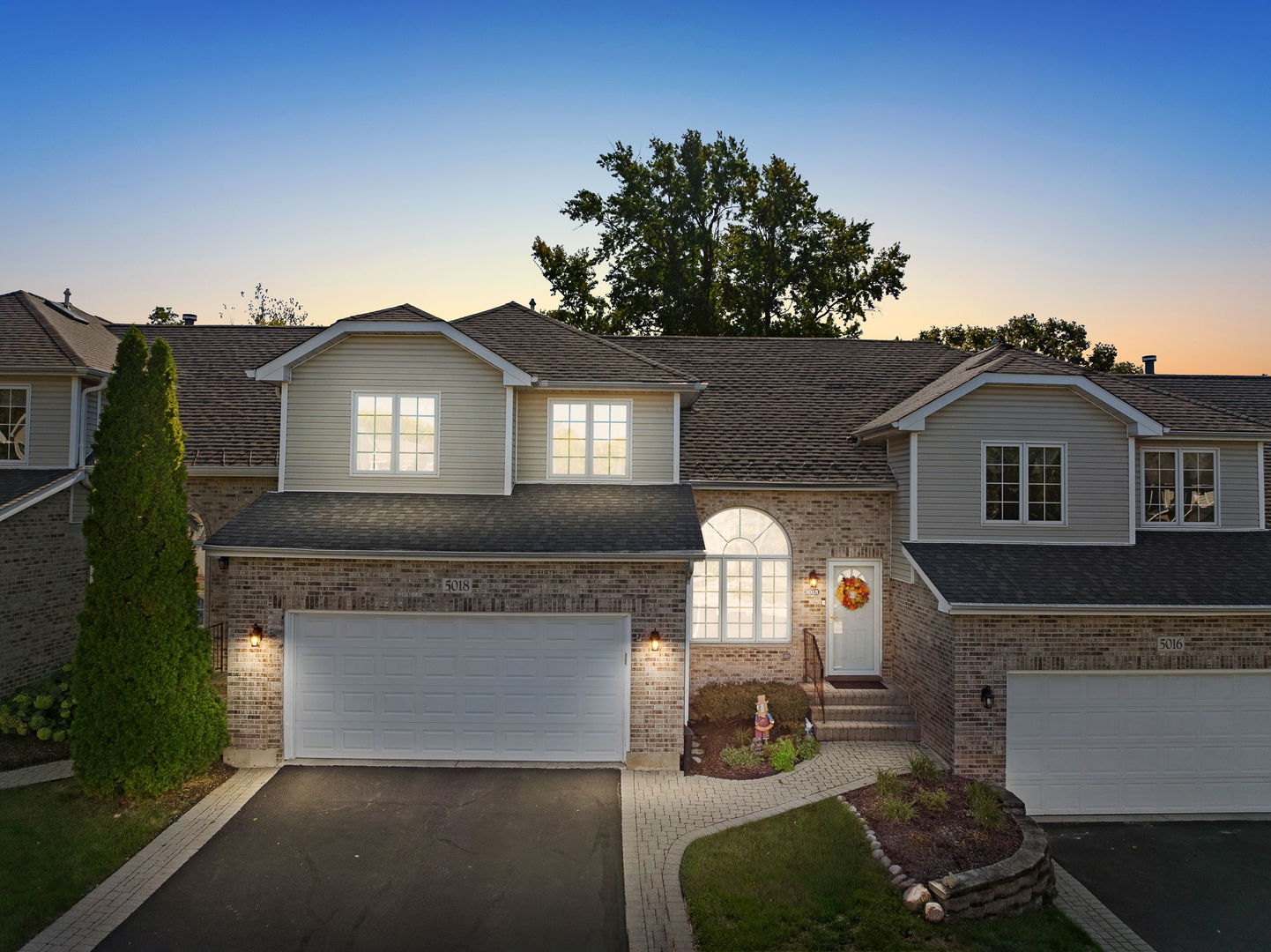 Two-story brick and beige-sided home with a two-car garage and arched entry window at dusk.