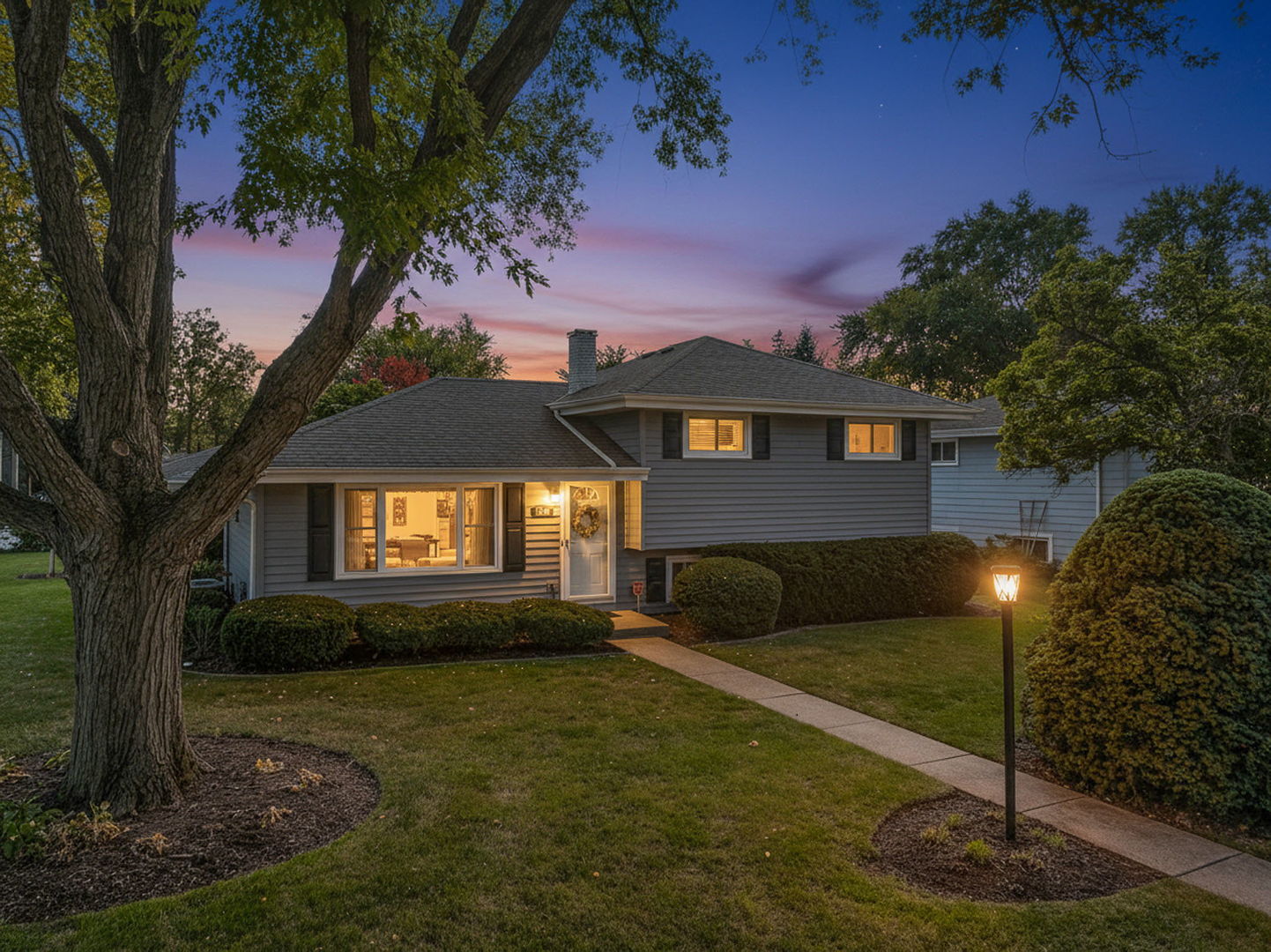 Gray two-story home at dusk, framed by a large tree, with warmly lit windows and a walkway to the front door.