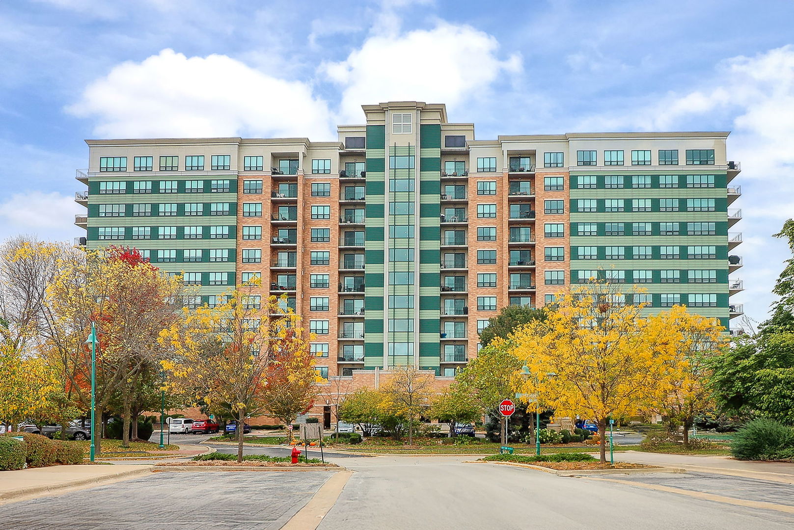Multistory condominium with green siding and brick sections, framed by yellow autumn trees.