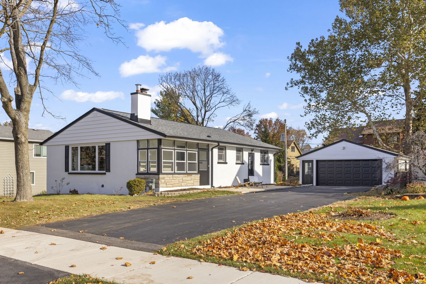 Single-story white house with dark-trimmed windows, detached garage, and leaf-covered autumn lawn.