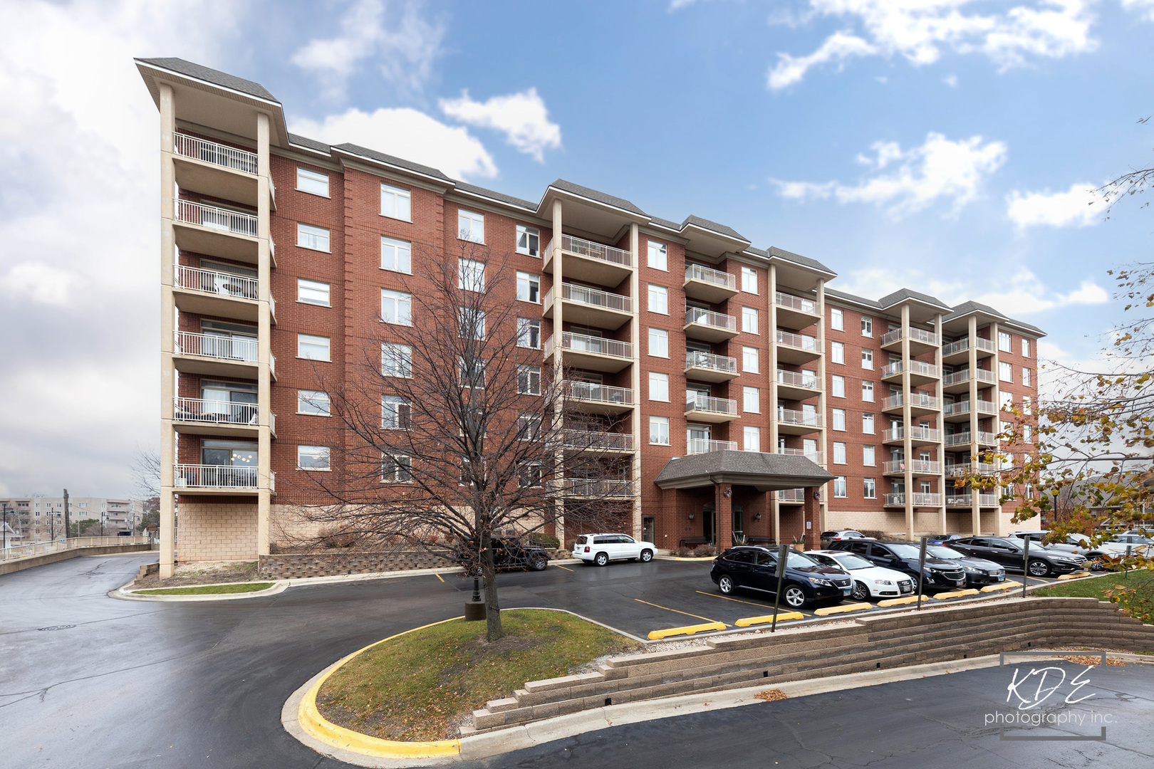Red-brick condominium building with cream balconies, a covered entrance, and parked cars.