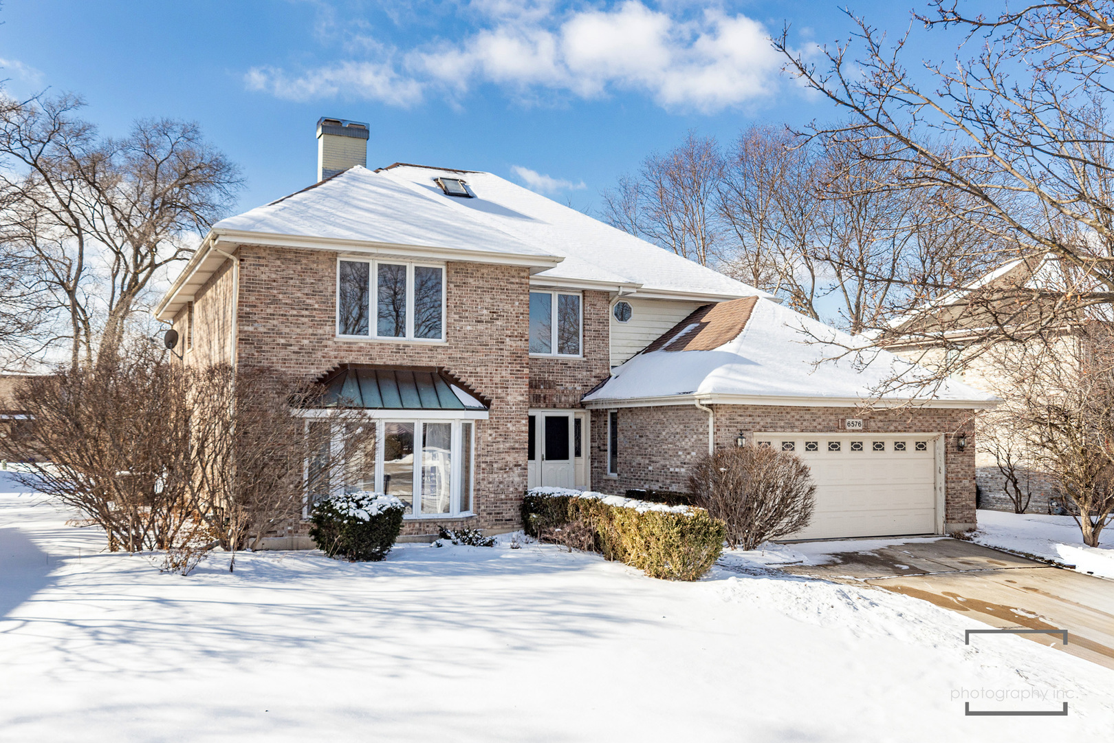Two-story brick house with an attached garage, snow-covered roof and yard, and leafless trees.
