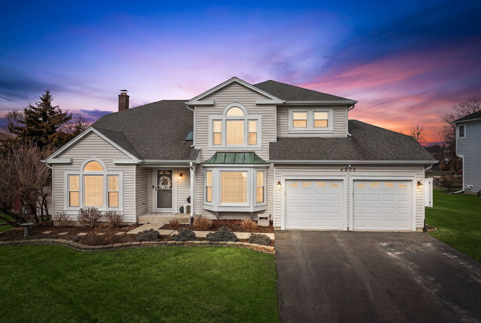 Two-story gray-sided home with a two-car garage and illuminated windows at dusk.