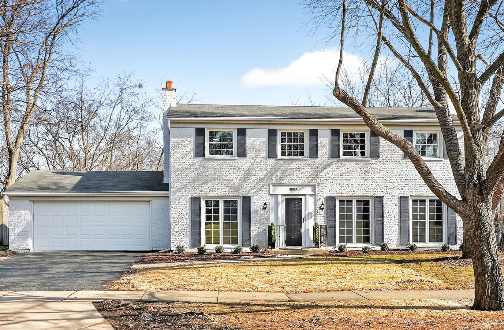 Two-story white brick house with dark shutters, attached garage, and leafless trees in the front yard.