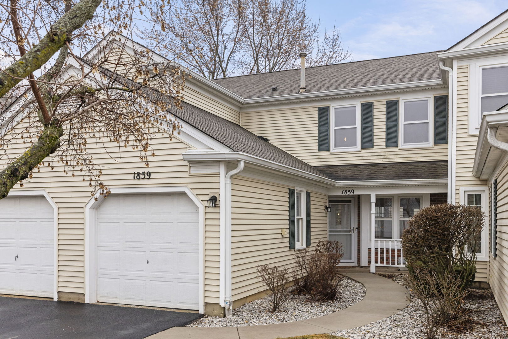 Two-story beige-sided home with white garage doors, green shutters, and a covered entry.