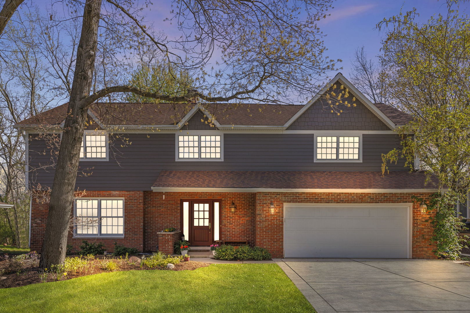 Two-story brick and dark-sided home with an attached garage and mature tree in front.