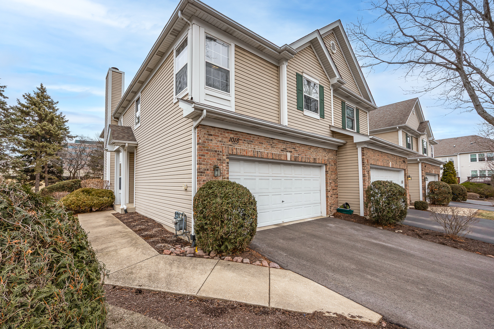 Two-story beige townhouse with brick garage surround, green shutters, and shrubs along the front walkway.