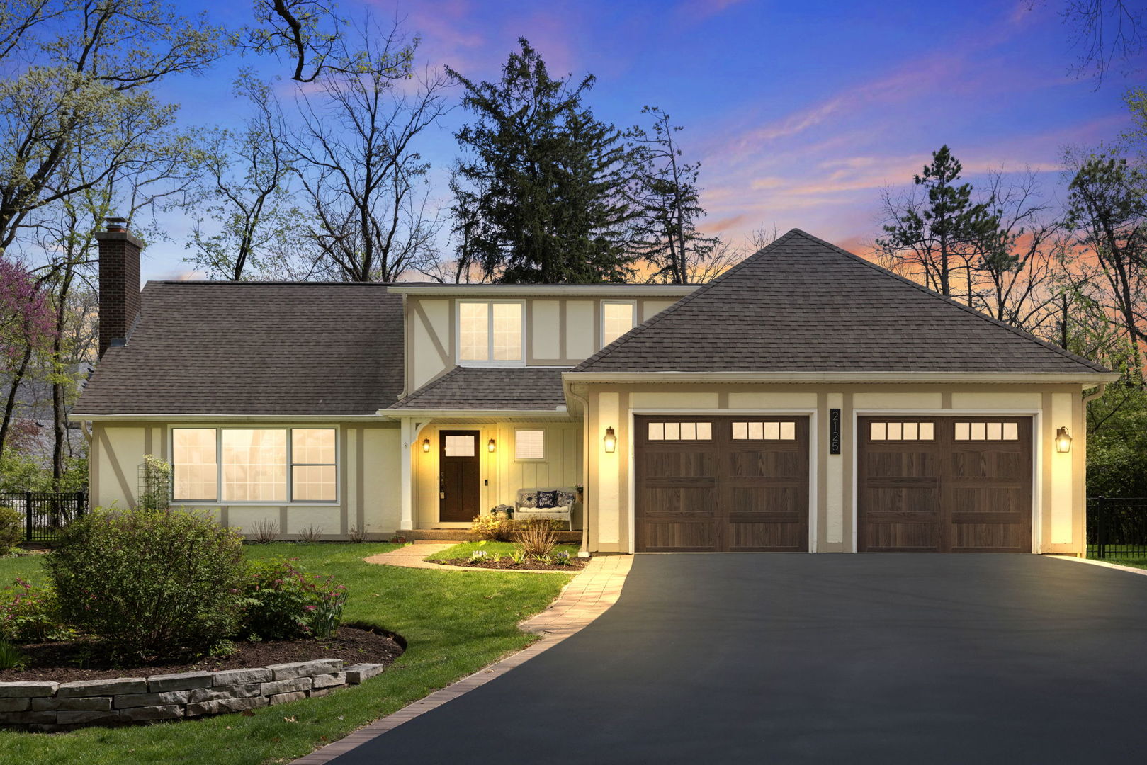 Suburban home with a two-car garage, landscaped front lawn, and warm lights at dusk.
