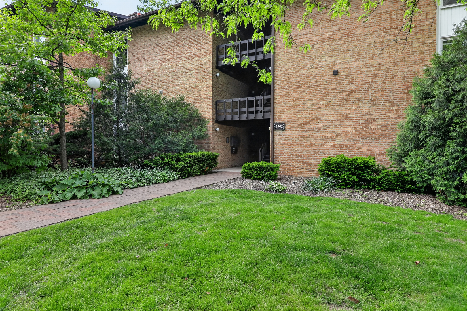 Brick condominium entrance with a paved walkway, landscaped shrubs, and a broad green lawn.