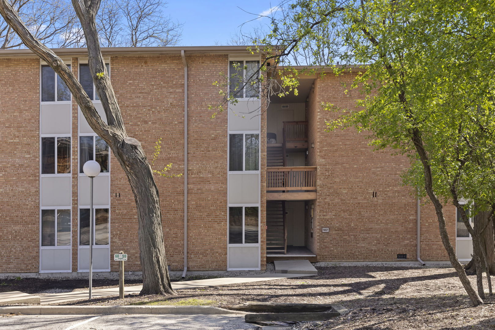 Brick apartment building with a recessed entrance, exterior stairs, and mature trees.