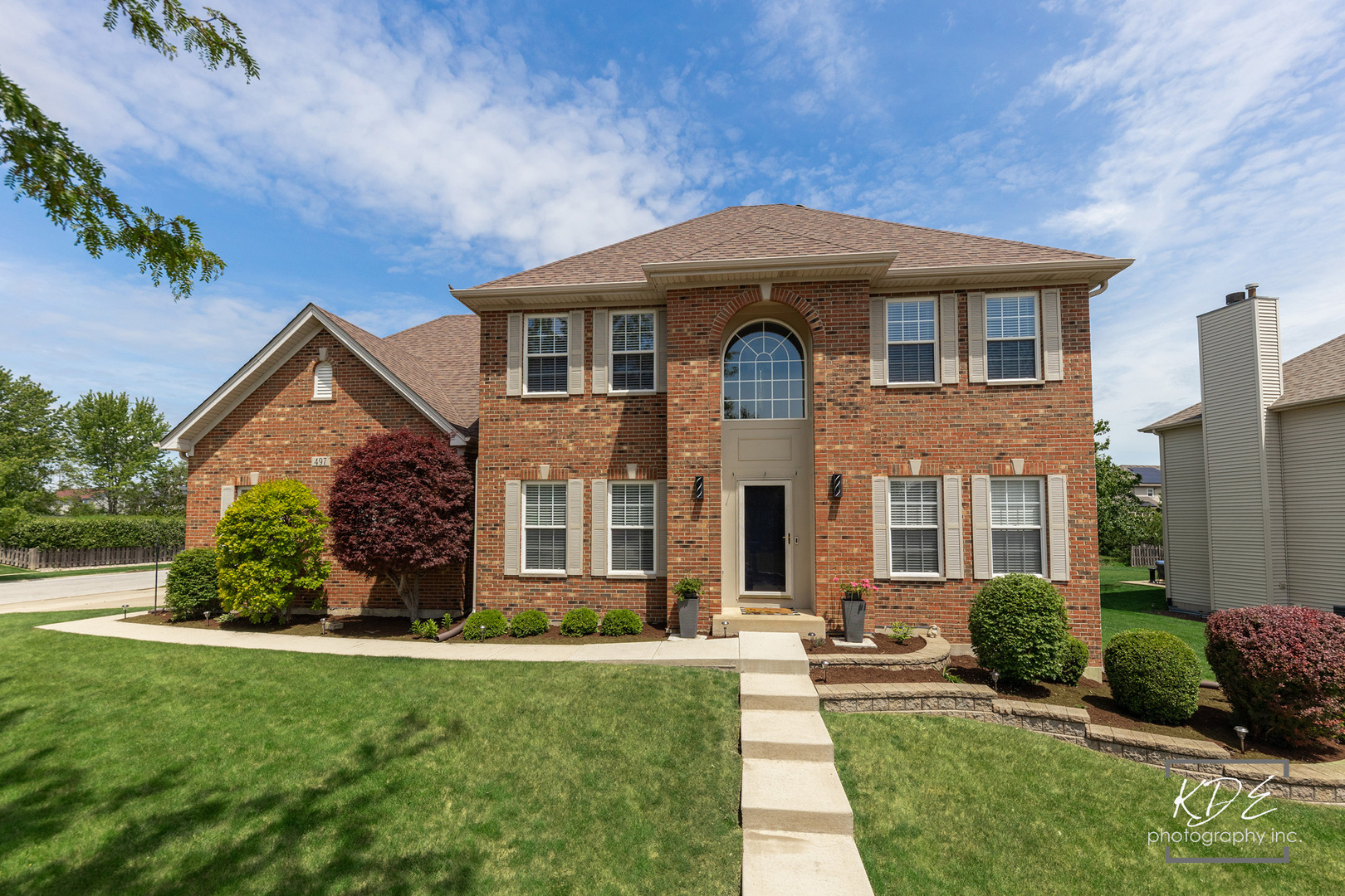 Two-story red-brick home with an arched entry window, landscaped lawn, and walkway.