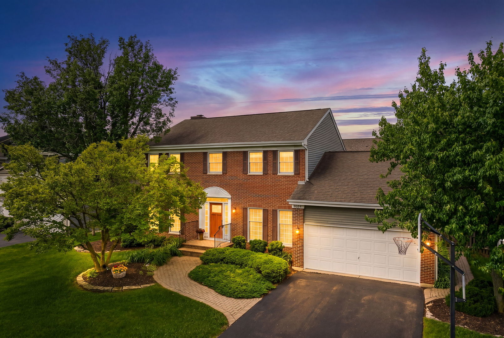 Two-story brick home at dusk with a landscaped yard, attached garage, and basketball hoop.