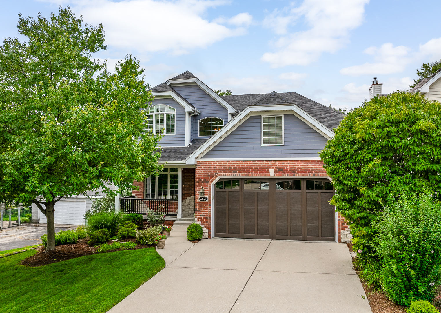 Two-story home with gray siding, red brick, a wide garage, and leafy landscaping.