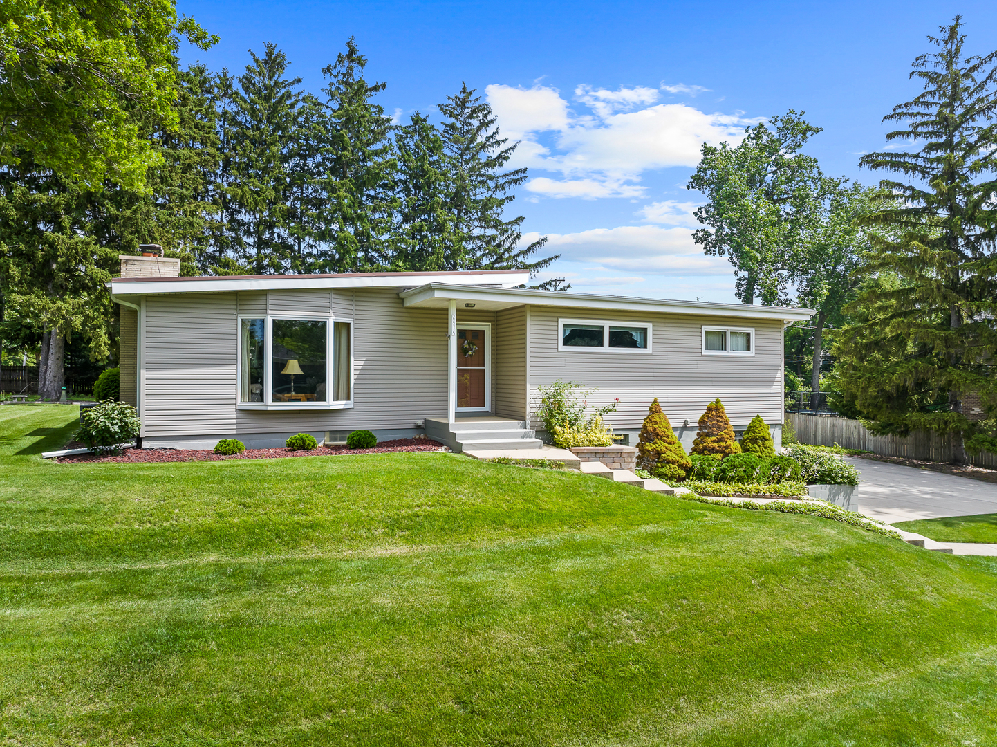 Single-story ranch home with gray siding, landscaped shrubs, and a broad green lawn.