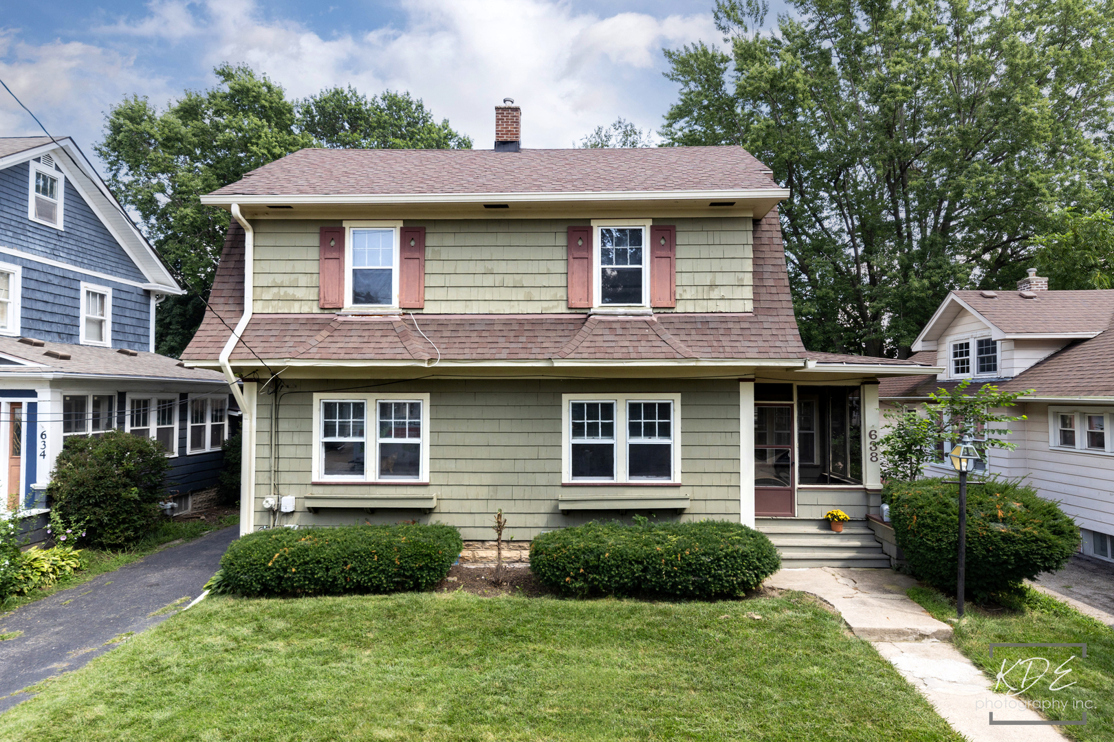 Two-story green-shingled house with red-brown shutters, a covered entry, and landscaped front lawn.