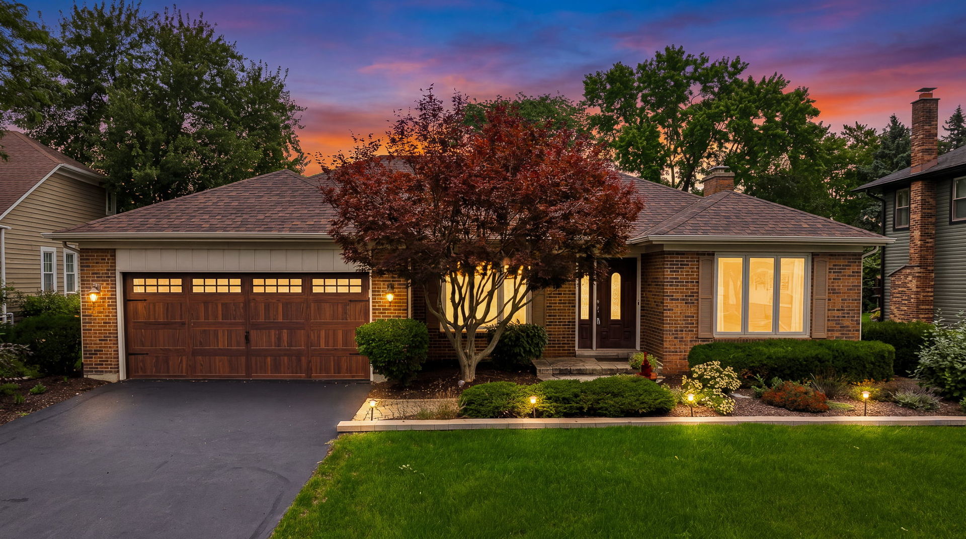 Single-story brick house at dusk with a wide wooden garage door and landscaped front garden.