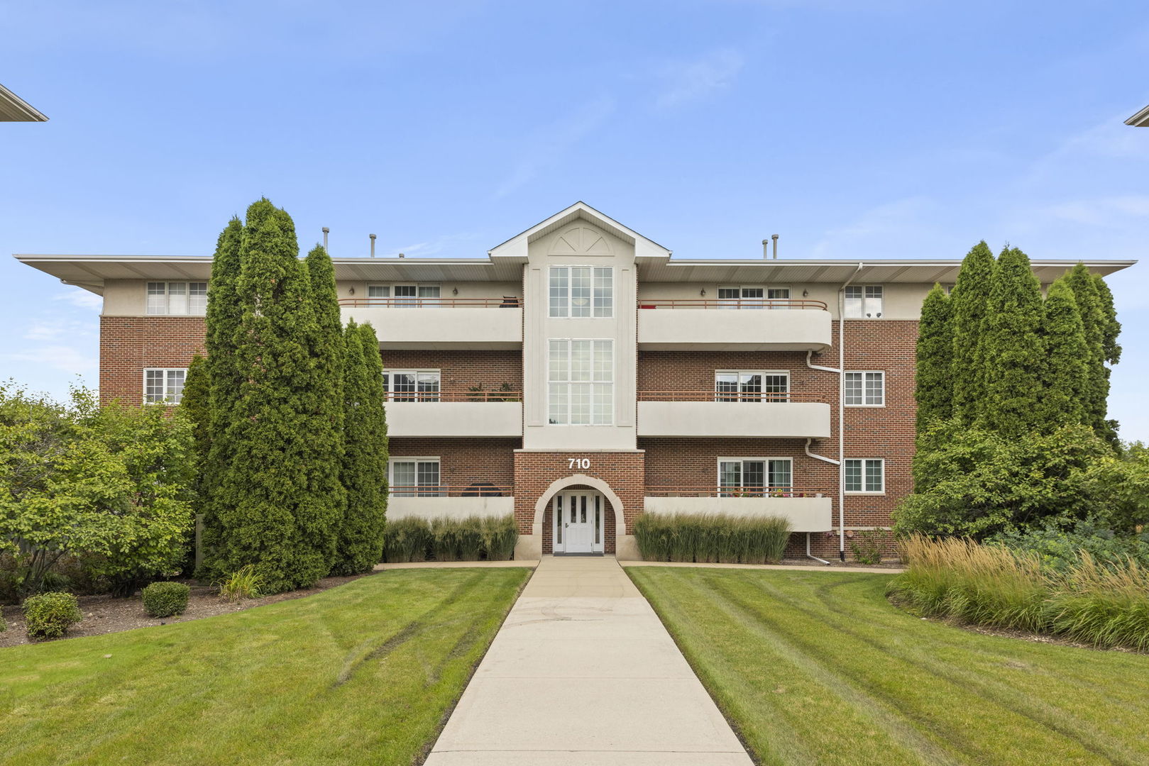 Three-story brick condominium building with cream balconies, an arched entrance, and a walkway across the lawn.